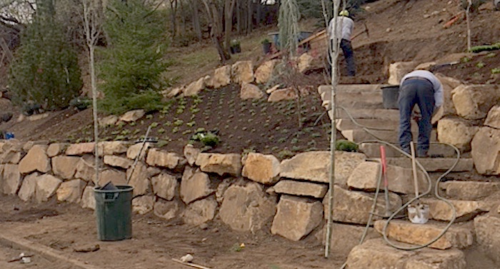 boulder retaining wall and stone steps being built and two landscaping workers are in the scene